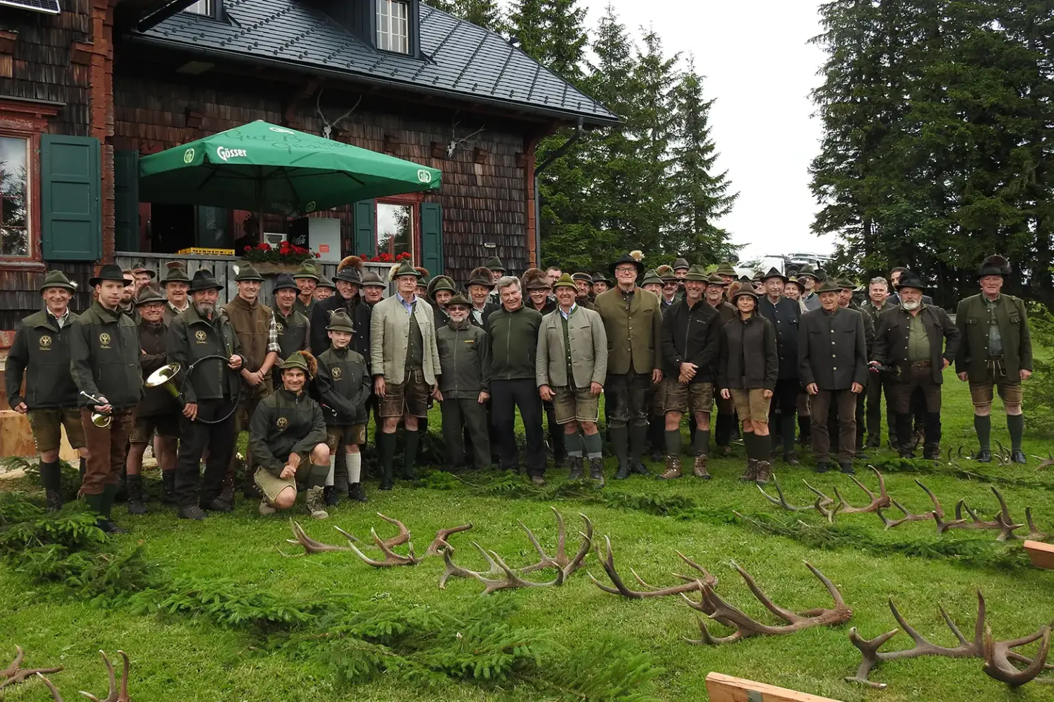 Eine große Gruppe von Personen in jagdlicher Kleidung steht auf einer Wiese vor einer Reihe von Geweihen