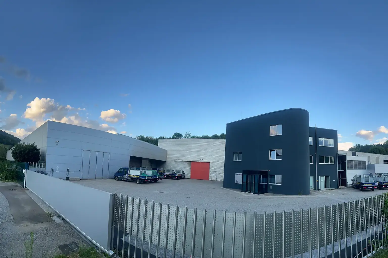 Das Gelände eines Gewerbebetriebes mit Produktionshalle und Bürogebäude hinter verschlossenem Tor, auf dem Parkplatz einige Pritschenwagen