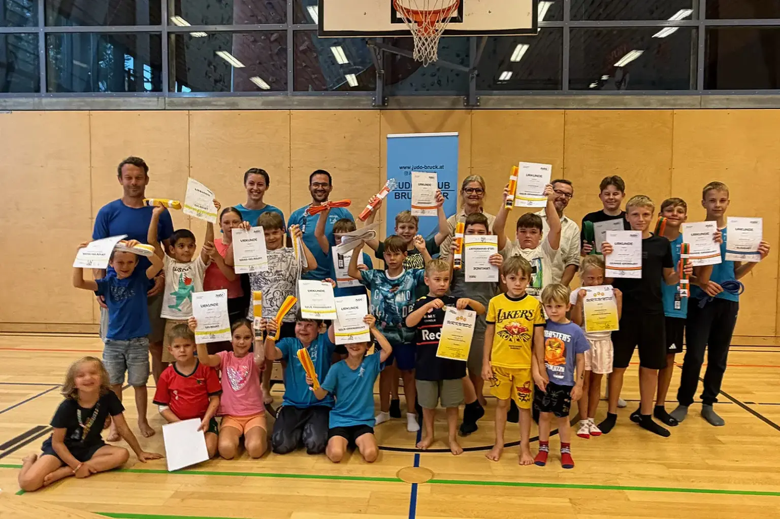 Feierliche Gürtelübergabe beim Judo-Club Bruck Eine große Gruppe von Kindern und mehreren Erwachsenen steht in einer Turnhalle für ein Gruppenfoto. Viele Kinder halten Urkunden und orange-weiße Gürtel in die Höhe. Im Hintergrund sind ein Basketballkorb und ein Banner des Judoclubs Bruck zu sehen.