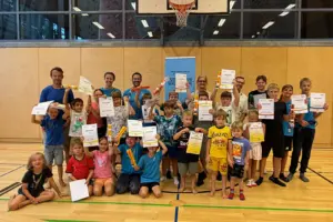 Eine große Gruppe von Kindern und mehreren Erwachsenen steht in einer Turnhalle für ein Gruppenfoto. Viele Kinder halten Urkunden und orange-weiße Gürtel in die Höhe. Im Hintergrund sind ein Basketballkorb und ein Banner des Judoclubs Bruck zu sehen.