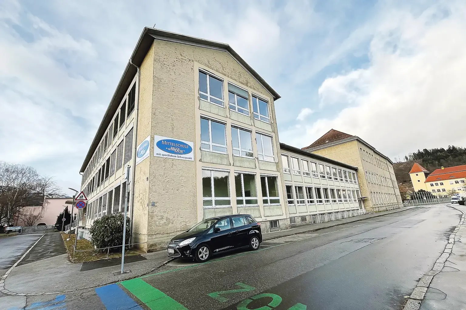 Ein dreigeschoßiges Gebäude, darauf eine Tafel mit der Aufschrift "Mittelschule Bruck an der Mur"