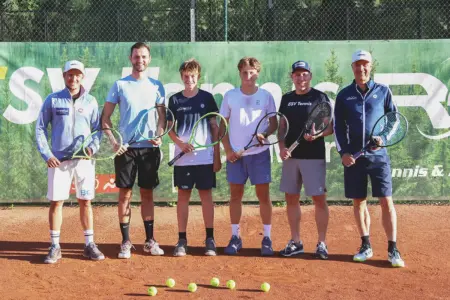 Tennisstars von morgen schlagen in Bruck auf Sechs Tennisspieler unterschiedlichen Alters stehen mit Tennisschlägern auf einem Sandplatz für ein Gruppenfoto. Vor ihnen liegen mehrere Tennisbälle, im Hintergrund ist ein Werbebanner am Zaun der Tennisanlage zu sehen.