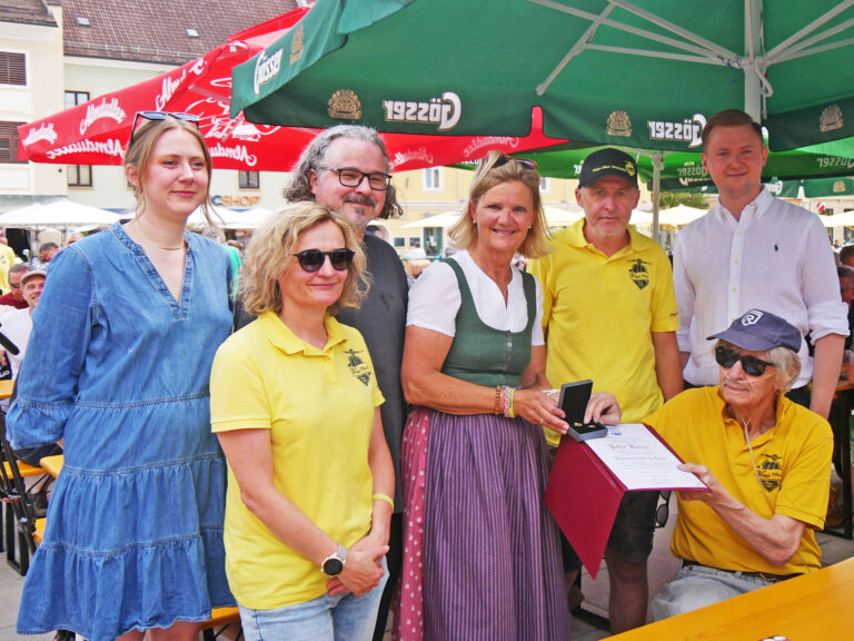 Sechs stehende Personen überreichen eine Auszeichnung an einen sitzenden Mann