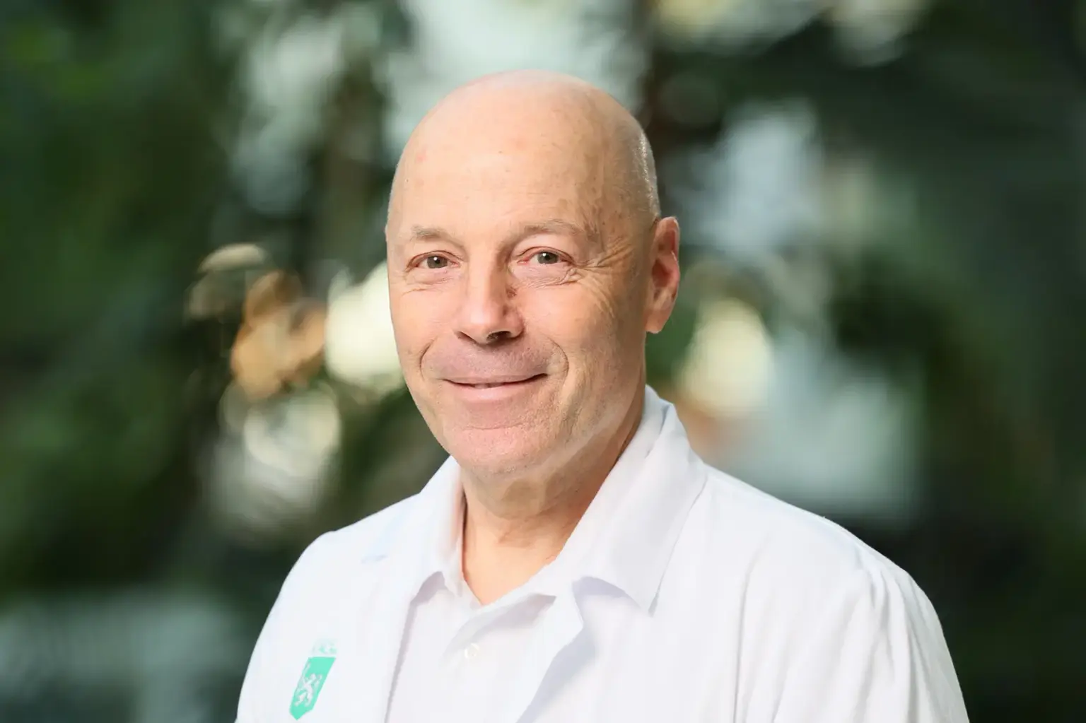 Porträt eines lächelnden, glatzköpfigen Mannes in einem weißen Hemd. Er blickt direkt in die Kamera, der Hintergrund ist unscharf und zeigt grüne Pflanzen in einem hellen Innenraum.