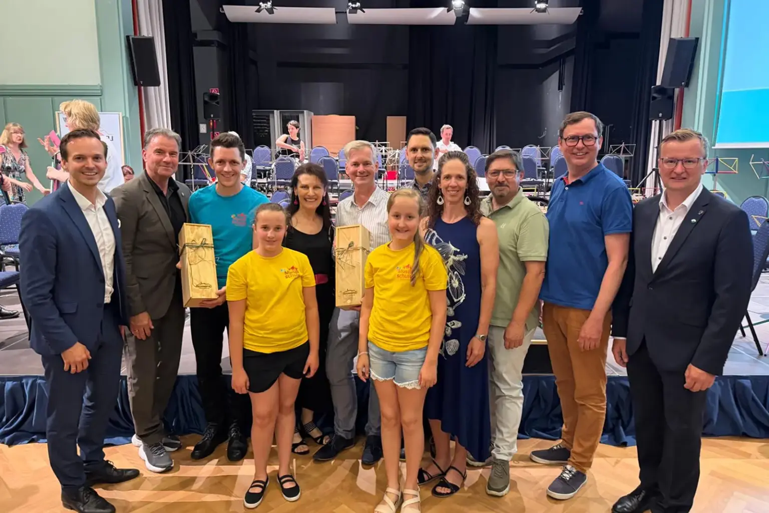 Eine Gruppe aus Erwachsenen und zwei Kindern steht in einem Veranstaltungssaal vor einer Bühne mit Orchesterstühlen. Zwei Personen halten Holzgeschenke in den Händen, die Kinder tragen gelbe T-Shirts der Musikschule und lächeln in die Kamera.
