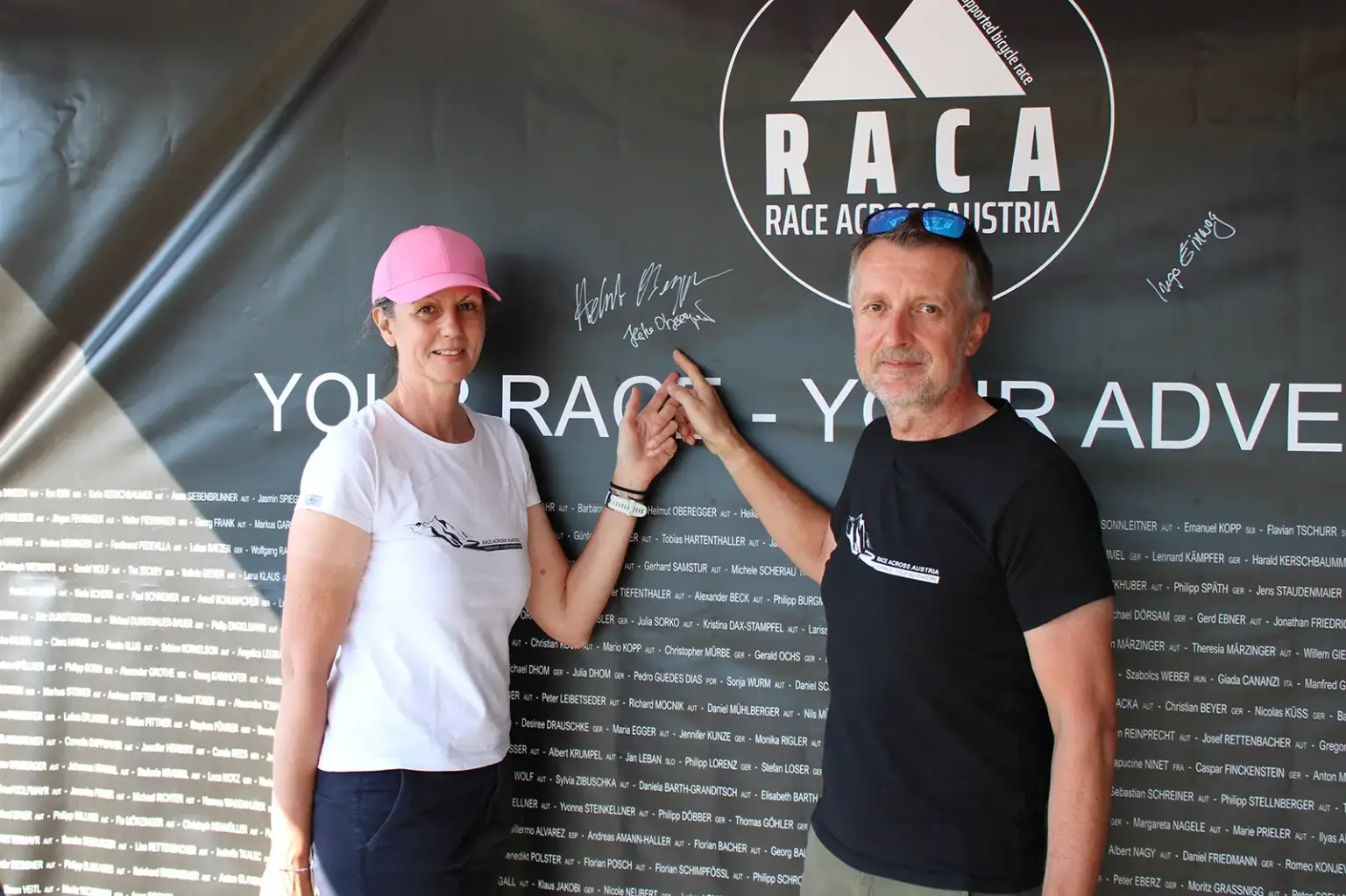 Eine Frau mit rosa Kappe und ein Mann in schwarzem T-Shirt stehen vor einer großen Sponsorentafel der Race Across Austria und zeigen auf eine Unterschrift.
