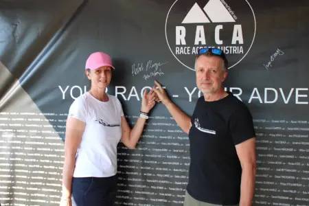 In 81 Stunden durch Österreich Eine Frau mit rosa Kappe und ein Mann in schwarzem T-Shirt stehen vor einer großen Sponsorentafel der Race Across Austria und zeigen auf eine Unterschrift.