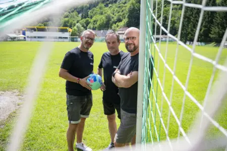 Die Rundschau bleibt am Ball Drei Männer stehen auf einem Fußballplatz neben einem Tor und halten gemeinsam einen Fußball. Im Hintergrund sind das Spielfeld, ein Vereinsgebäude und bewaldete Hügel zu sehen.
