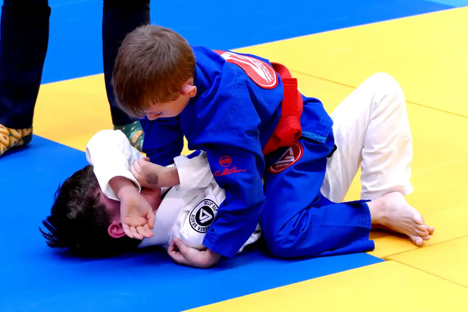 30 Medaillen für Kapfenberger BJJ-Kämpfer Zwei Kinder in blauen und weißen Brazilian-Jiu-Jitsu-Anzügen kämpfen auf einer gelb-blauen Matte. Das Kind im blauen Anzug befindet sich in der Oberlage und kontrolliert den Gegner am Boden.
