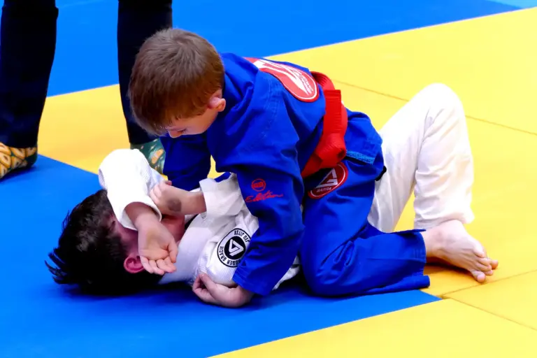 30 Medaillen für Kapfenberger BJJ-Kämpfer Zwei Kinder in blauen und weißen Brazilian-Jiu-Jitsu-Anzügen kämpfen auf einer gelb-blauen Matte. Das Kind im blauen Anzug befindet sich in der Oberlage und kontrolliert den Gegner am Boden.