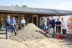 Acht Personen stehen mit roten Spaten rund um einen Sandhaufen vor dem Gebäude des Tierheims Kapfenberg und markieren gemeinsam den symbolischen Spatenstich für den Umbau. Im Hintergrund sind Bauzäune, das sanierungsbedürftige Gebäude und Baustelleneinrichtungen zu sehen.