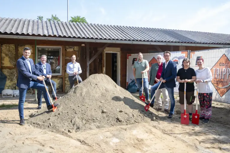 Spatenstich für neues Hundehaus im Kapfenberger Tierheim Acht Personen stehen mit roten Spaten rund um einen Sandhaufen vor dem Gebäude des Tierheims Kapfenberg und markieren gemeinsam den symbolischen Spatenstich für den Umbau. Im Hintergrund sind Bauzäune, das sanierungsbedürftige Gebäude und Baustelleneinrichtungen zu sehen.
