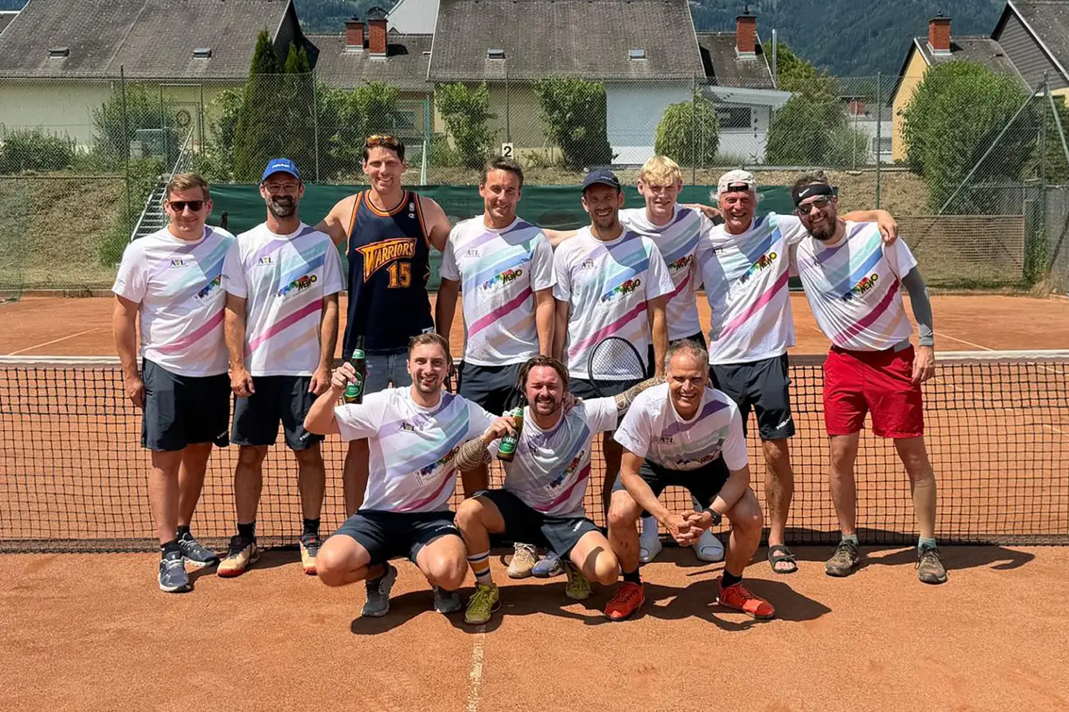 Elf Männer posieren nach einem Tennisturnier auf einem Sandplatz vor dem Netz. Die meisten tragen weiße Mannschaftsshirts, einige halten Tennisschläger oder Getränkeflaschen in der Hand. Im Hintergrund sind Wohnhäuser und bewaldete Hänge zu sehen.