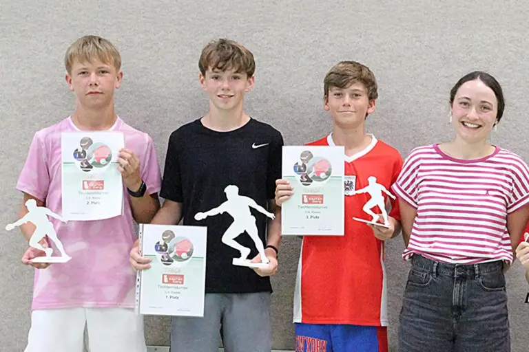 Gymnasium Kapfenberg: Schulmeister im Tischtennis Vier Jugendliche stehen nebeneinander vor einer hellen Wand. Drei halten Urkunden und stilisierte Tischtennis-Trophäen in den Händen, eine Jugendliche lächelt ohne Auszeichnung in die Kamera.
