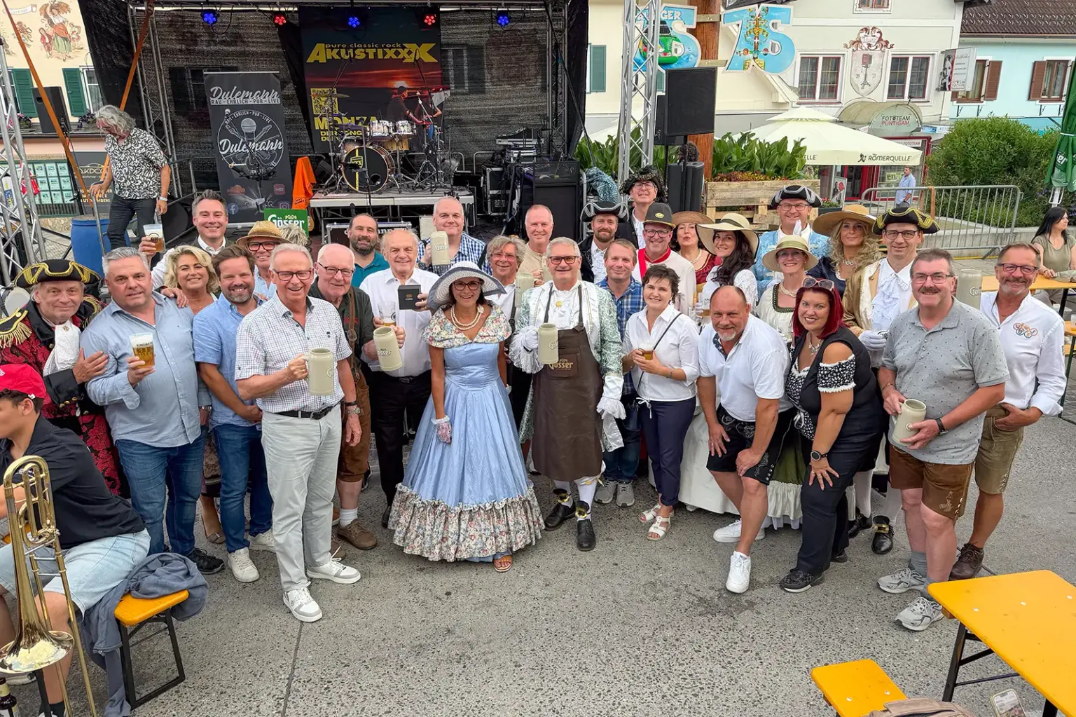 Rund 30 Teilnehmerinnen und Teilnehmer versammeln sich beim Kindberger Kirtag vor einer Bühne zu einem Gruppenfoto. Viele tragen historische Kostüme, Trachten oder festliche Kleidung und halten Bierkrüge in die Kamera. Im Hintergrund sind die Bühne, Musikinstrumente und die Kirtagskulisse am Hauptplatz zu sehen.
