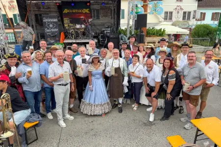 Rund 30 Teilnehmerinnen und Teilnehmer versammeln sich beim Kindberger Kirtag vor einer Bühne zu einem Gruppenfoto. Viele tragen historische Kostüme, Trachten oder festliche Kleidung und halten Bierkrüge in die Kamera. Im Hintergrund sind die Bühne, Musikinstrumente und die Kirtagskulisse am Hauptplatz zu sehen.