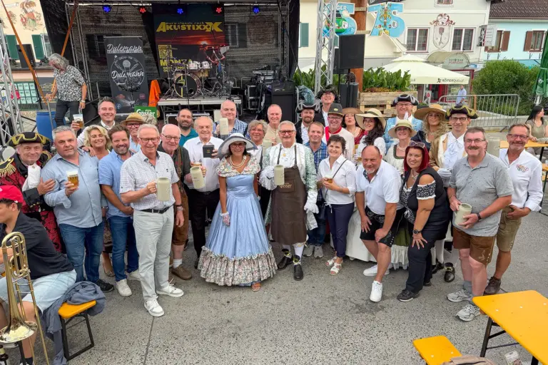 Rund 30 Teilnehmerinnen und Teilnehmer versammeln sich beim Kindberger Kirtag vor einer Bühne zu einem Gruppenfoto. Viele tragen historische Kostüme, Trachten oder festliche Kleidung und halten Bierkrüge in die Kamera. Im Hintergrund sind die Bühne, Musikinstrumente und die Kirtagskulisse am Hauptplatz zu sehen.