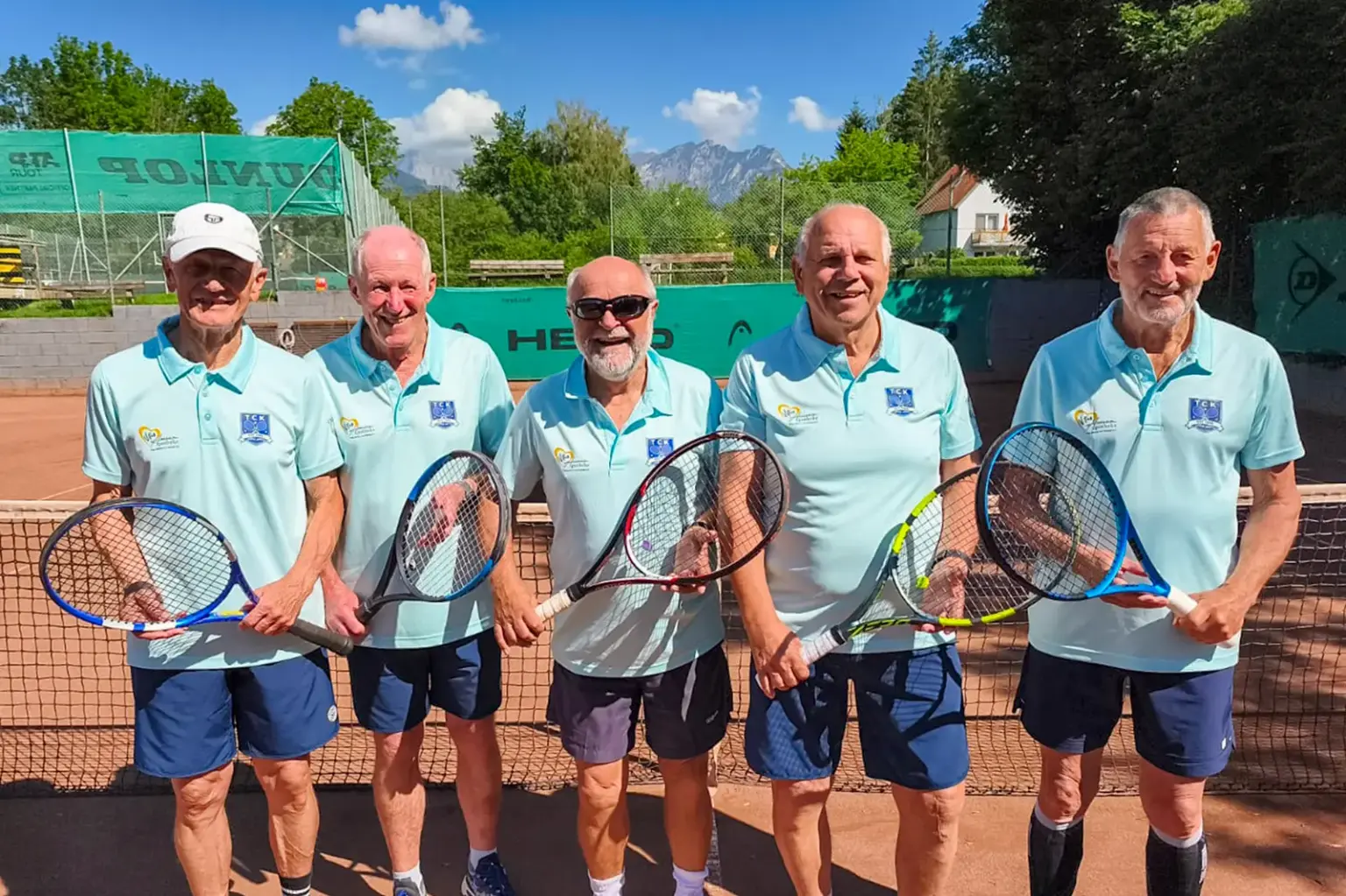 Fünf ältere Männer stehen mit Tennisschlägern auf einem Sandplatz vor dem Netz. Sie tragen hellblaue Poloshirts und lächeln in die Kamera. Im Hintergrund sind Tennisplätze, Bäume und Berge zu sehen.