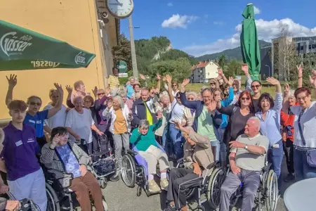 Eine Gruppe von älteren Personen, zum Teil in Rollstühlen, steht winkend auf einem Platz vor einem Haus