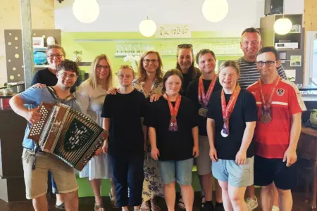Eine Gruppe aus Jugendlichen und Erwachsenen steht gemeinsam in einem Café und lächelt in die Kamera. Mehrere Personen tragen Medaillen um den Hals, eine Person im Vordergrund hält eine steirische Harmonika.
