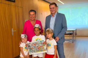 Drei Kindergartenkinder mit Eulen-Kronen präsentieren gemeinsam ein Wimmelbuch und werden dabei von einer Frau in pinker Bluse sowie einem Mann im hellblauen Anzug begleitet. Die Aufnahme entstand in einem hellen Innenraum mit Holzvertäfelung.