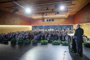 Ein Mann steht mit dem Rücken zum Betrachter vor nahezu vollbesetzten Sesselreihen in einem Veranstaltungssaal.