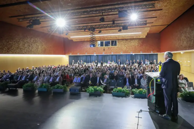 Ein Mann steht mit dem Rücken zum Betrachter vor nahezu vollbesetzten Sesselreihen in einem Veranstaltungssaal.