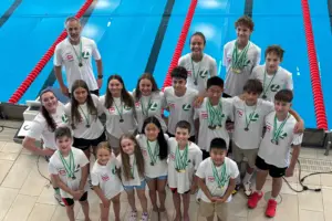 Gruppenfoto eines Schwimmteams am Beckenrand eines Hallenbads. Rund 20 Kinder und Jugendliche sowie zwei Trainer tragen weiße Vereins-Shirts und präsentieren zahlreiche Medaillen nach einem erfolgreichen Wettkampf. Im Hintergrund sind die Schwimmbahnen mit roten Leinen zu sehen.
