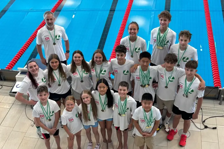 Heimische Schwimmer sind steirische Meister Gruppenfoto eines Schwimmteams am Beckenrand eines Hallenbads. Rund 20 Kinder und Jugendliche sowie zwei Trainer tragen weiße Vereins-Shirts und präsentieren zahlreiche Medaillen nach einem erfolgreichen Wettkampf. Im Hintergrund sind die Schwimmbahnen mit roten Leinen zu sehen.