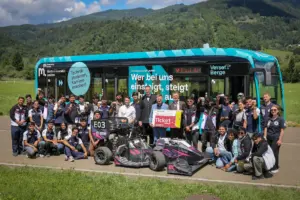 Eine große Gruppe von Personen posiert mit einem Rennwagen vor einem Bus