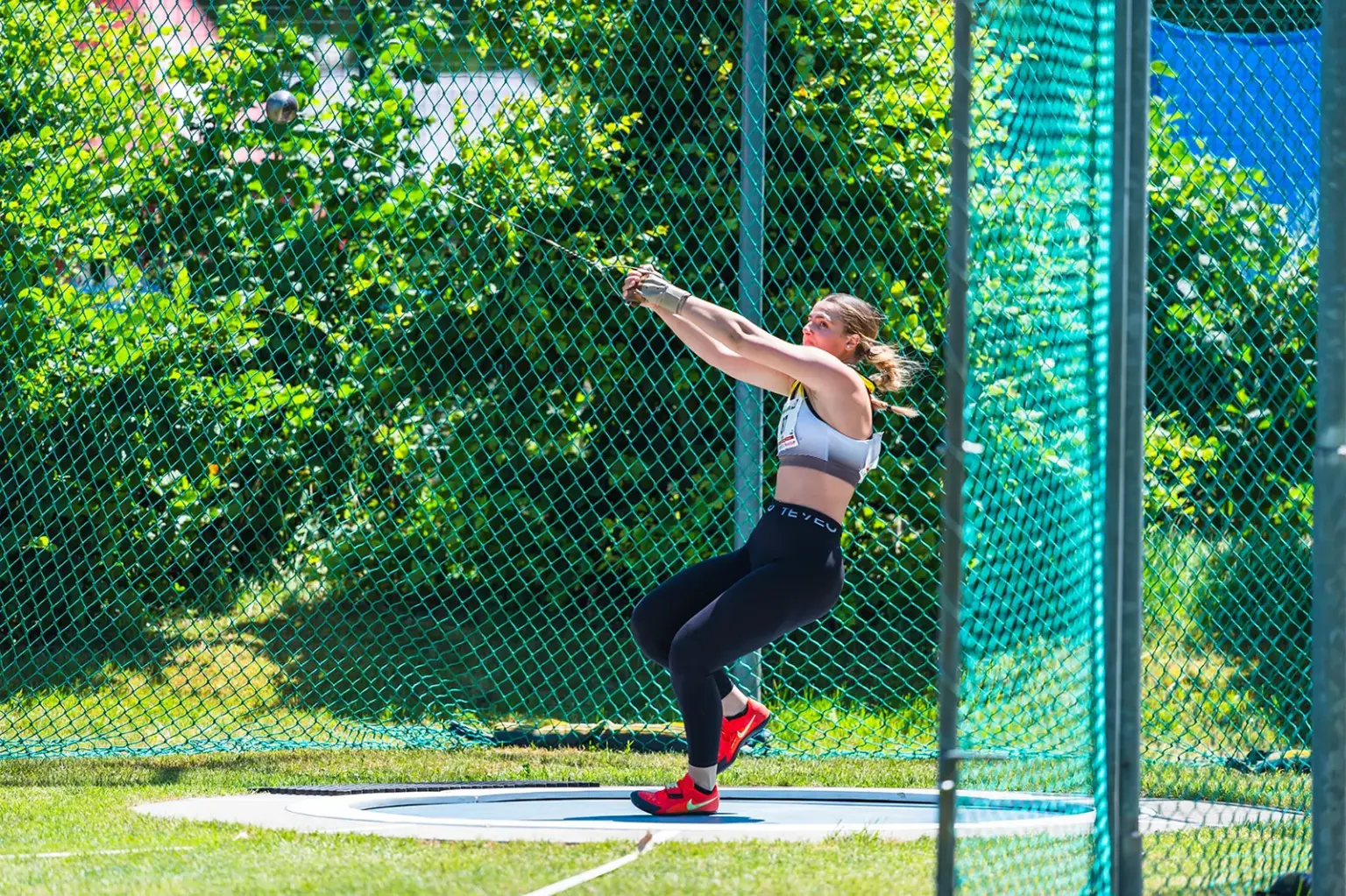 Eine Leichtathletin wirbelt im Wurfkäfig den Hammer durch die Luft. Sie steht im Wurfkreis, trägt Sportbekleidung und rote Schuhe, während das Schutznetz und grüne Bäume den Hintergrund bilden.