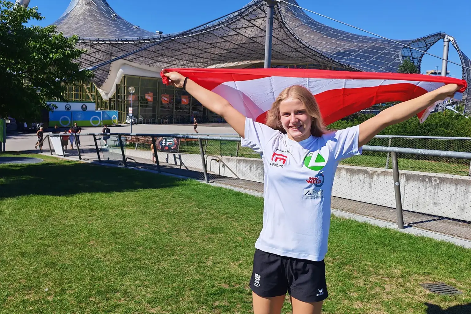 Eine junge Sportlerin steht vor dem Münchner Olympiastadion und hält eine rot-weiß-rote Österreich-Flagge mit ausgestreckten Armen über den Schultern. Sie trägt ein weißes Vereins-Shirt und lächelt in die Kamera.