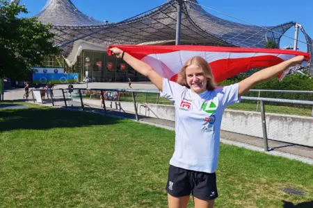Eine junge Sportlerin steht vor dem Münchner Olympiastadion und hält eine rot-weiß-rote Österreich-Flagge mit ausgestreckten Armen über den Schultern. Sie trägt ein weißes Vereins-Shirt und lächelt in die Kamera.