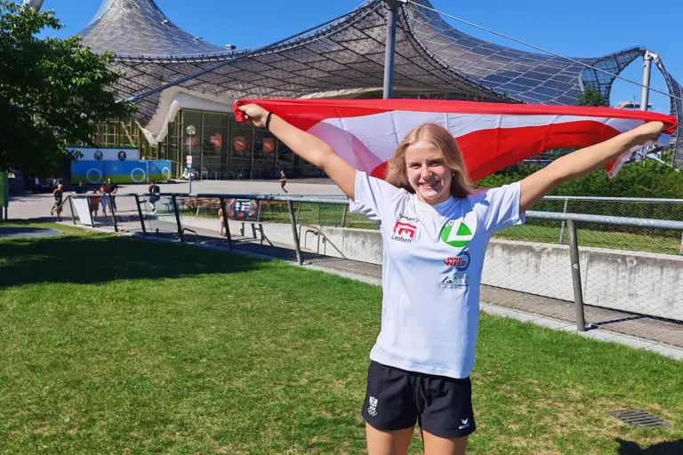 Eine junge Sportlerin steht vor dem Münchner Olympiastadion und hält eine rot-weiß-rote Österreich-Flagge mit ausgestreckten Armen über den Schultern. Sie trägt ein weißes Vereins-Shirt und lächelt in die Kamera.