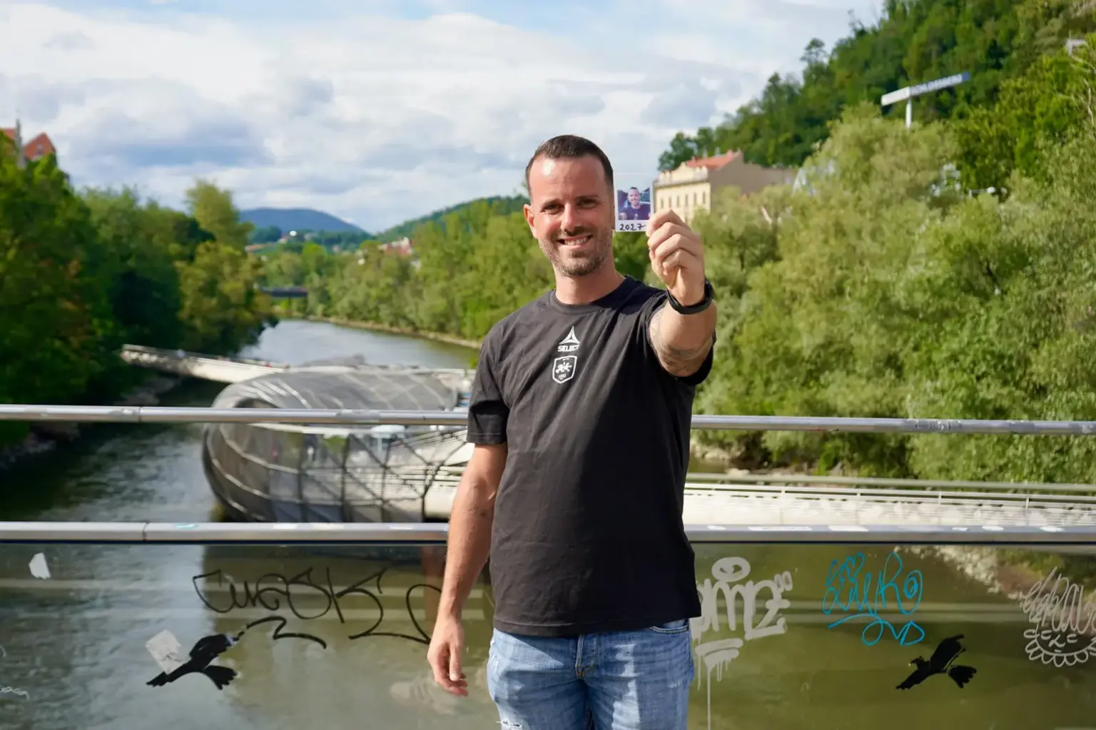 Ein Mann steht auf einer Brücke über der Mur in Graz und hält ein kleines Passfoto in die Kamera. Im Hintergrund sind die Murinsel, die Uferpromenade und bewaldete Hänge zu sehen.