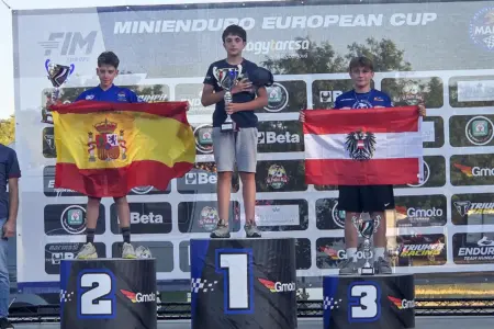 Zwei Stockerlplätze für jungen Trofaiacher Endurofahrer Drei Jugendliche stehen mit Pokalen auf einem Siegerpodest vor einer Sponsorentafel mit der Aufschrift „Minienduro European Cup“. Der Zweitplatzierte hält eine spanische Flagge, der Drittplatzierte eine österreichische Flagge. Der Erstplatzierte steht in der Mitte auf Platz eins und hält einen Pokal sowie eine Kappe in den Händen.