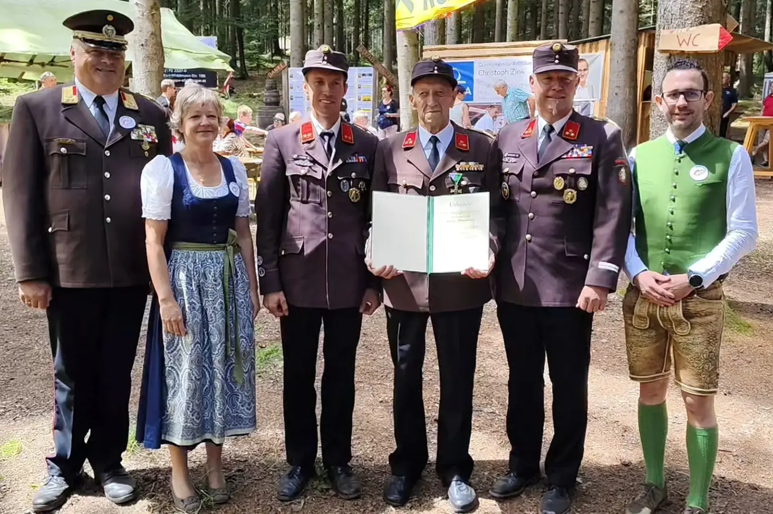 Sechs Personen stehen bei einer Feier im Wald für ein Gruppenfoto zusammen. Drei Mitglieder der Freiwilligen Feuerwehr tragen Ausgehuniformen, der Mann in der Mitte hält eine geöffnete Ehrenurkunde. Flankiert werden sie von einer Frau im Dirndl, einem Feuerwehrfunktionär sowie einem Mann in steirischer Tracht. Im Hintergrund sind Feststände und Besucher zu sehen.