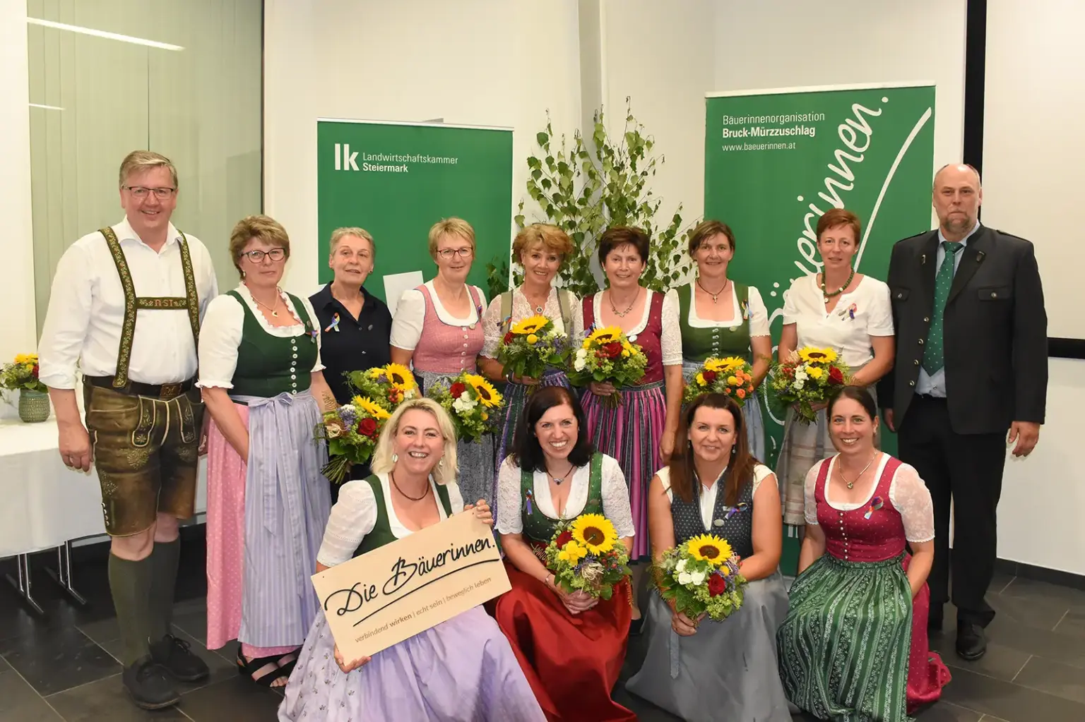 Eine Gruppe von 14 Personen steht und kniet für ein Gruppenfoto in einem Veranstaltungsraum. Mehrere Frauen tragen Dirndl und halten Blumensträuße in den Händen. Im Hintergrund sind zwei grüne Roll-up-Banner sowie eine dekorative Birke zu sehen.