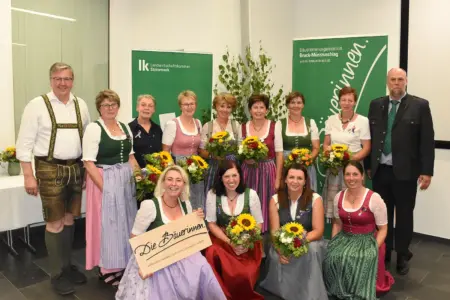 Heimische Bäuerinnen und Bauern wurden ausgezeichnet Eine Gruppe von 14 Personen steht und kniet für ein Gruppenfoto in einem Veranstaltungsraum. Mehrere Frauen tragen Dirndl und halten Blumensträuße in den Händen. Im Hintergrund sind zwei grüne Roll-up-Banner sowie eine dekorative Birke zu sehen.