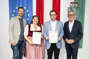 Vier Personen stehen vor den Fahnen der Europäischen Union, Österreichs und der Steiermark. Zwei Personen in der Mitte halten Urkunden in den Händen und blicken in die Kamera.