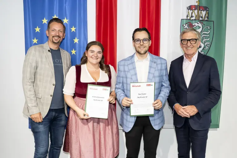 Vier Personen stehen vor den Fahnen der Europäischen Union, Österreichs und der Steiermark. Zwei Personen in der Mitte halten Urkunden in den Händen und blicken in die Kamera.