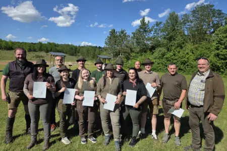 Eine Gruppe von Frauen und Männern steht auf einer Wiese vor einem Schießstand und hält Urkunden in die Kamera. Die Teilnehmenden tragen überwiegend jagdliche Kleidung und Hüte, im Hintergrund sind Bäume und ein blauer Himmel zu sehen.