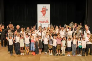 Schülerinnen und Schüler der Johann-Brahms-Musikschule Mürzzuschlag präsentieren gemeinsam mit ihren Lehrkräften auf einer Bühne ihre Instrumente. Die große Gruppe hält unter anderem Geigen, Trompeten, Hörner, Tuben und Klarinetten in die Höhe. Im Hintergrund ist ein Banner der Musikschule zu sehen.
