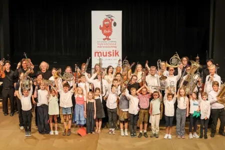 Abschlusskonzerte der Musikschule Mürzzuschlag Schülerinnen und Schüler der Johann-Brahms-Musikschule Mürzzuschlag präsentieren gemeinsam mit ihren Lehrkräften auf einer Bühne ihre Instrumente. Die große Gruppe hält unter anderem Geigen, Trompeten, Hörner, Tuben und Klarinetten in die Höhe. Im Hintergrund ist ein Banner der Musikschule zu sehen.
