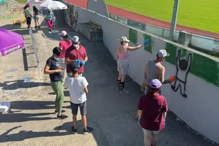 Mürzzuschlager Skatepark im neuen Design Mehrere Personen reinigen und übermalen Graffiti an einer Betonwand in einer Skateanlage. Einige tragen Handschuhe und arbeiten mit Farbrollen, andere stehen daneben und helfen.