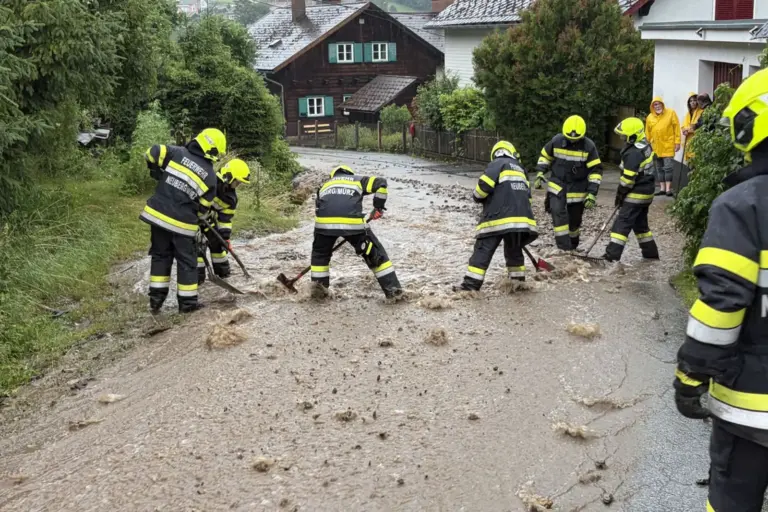 Unwettereinsätze im Mürztal Mehrere Feuerwehrleute räumen nach einem Unwetter mit Schaufeln und Rechen Schlamm und Geröll von einer überfluteten Straße in einem Wohngebiet. Im Hintergrund beobachten Anwohner die Aufräumarbeiten.