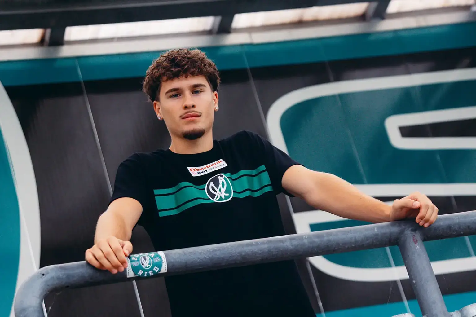 Ein junger Mann in schwarzem T-Shirt lehnt an einem Metallgeländer und blickt in die Kamera. Im Hintergrund ist eine Gebäudefassade mit großflächigen grünen und schwarzen Elementen zu sehen.