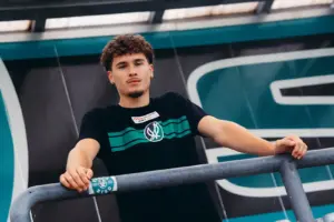 Pernegger Kicker wechselt zur SV Ried Ein junger Mann in schwarzem T-Shirt lehnt an einem Metallgeländer und blickt in die Kamera. Im Hintergrund ist eine Gebäudefassade mit großflächigen grünen und schwarzen Elementen zu sehen.