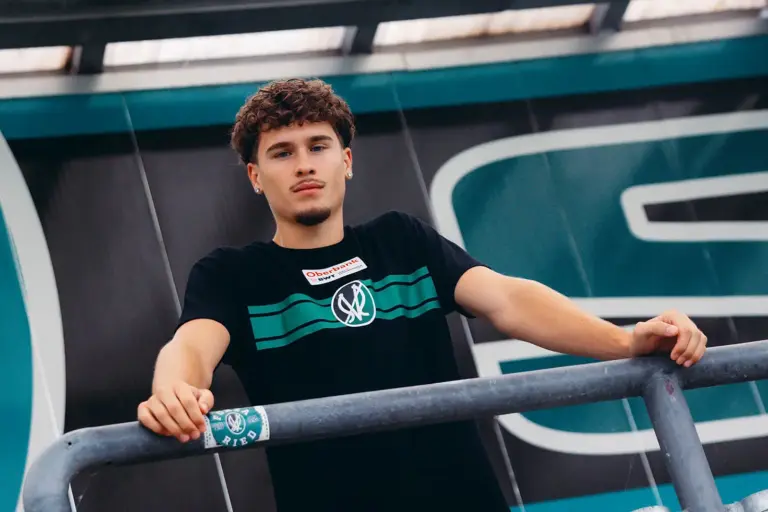 Pernegger Kicker wechselt zur SV Ried Ein junger Mann in schwarzem T-Shirt lehnt an einem Metallgeländer und blickt in die Kamera. Im Hintergrund ist eine Gebäudefassade mit großflächigen grünen und schwarzen Elementen zu sehen.