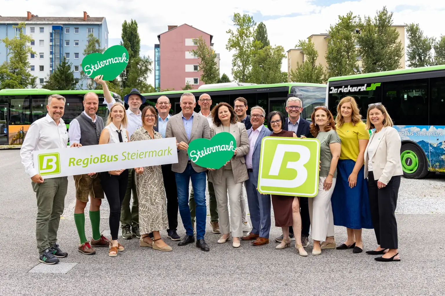 Schöner fahren: Steirische Regionen zieren Linienbusse Eine Gruppe von zirka 15 Menschen mit Schildern in Händen, im Hintergrund Linienbusse
